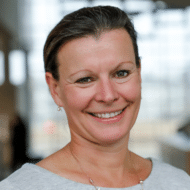 Porträt einer lächelnden Frau mit kurzen, grauen Haaren, in einem hellen Pullover, vor einem modernen Hintergrund.