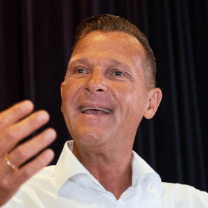 Lächelnder Mann in weißem Hemd, spricht mit erhobener Hand vor dunklem Hintergrund.