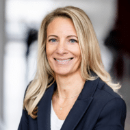 Lächelnde Frau mit langen blonden Haaren, trägt einen dunklen Blazer, Hintergrund unscharf.