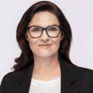 Porträt einer Frau mit langen, dunklen Haaren, Brille und schwarzem Blazer vor einem hellen Hintergrund.