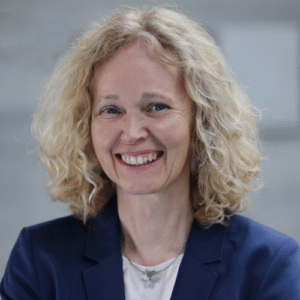 Lächelnde Frau mit lockigem, blondem Haar, trägt einen blauen Blazer, vor einem neutralen Hintergrund.