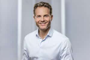 Mann mit kurzem, blondem Haar und lächelndem Gesicht, trägt ein weißes Hemd, vor einem neutralen Hintergrund.