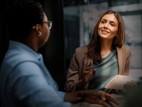 Zwei Personen in einem Gespräch, eine Frau mit Tablet lächelt, der andere sitzt mit Brille und hört aufmerksam zu.