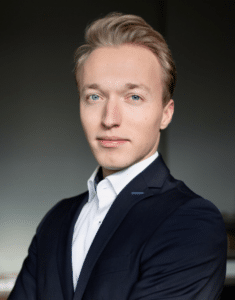 Junger Mann mit blonden Haaren, in einem schwarzen Anzug, schaut direkt in die Kamera mit neutralem Gesichtsausdruck.