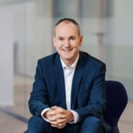 Mann in Anzug sitzt lächelnd auf einem Stuhl in einem modernen Büro mit Glaswänden.