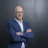 Mann mit Brille und Anzug steht mit verschränkten Armen vor einem dunklen Hintergrund.