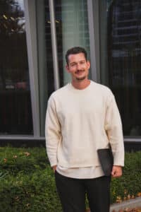 Mann mit kurzem Haar und Bart, trägt einen hellen Pullover und hält einen Laptop, steht vor modernen Fenstern und Pflanzen.
