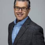 Porträt eines Mannes mit Brille, grauem Anzug und blauem Hemd, lächelnd vor einem neutralen Hintergrund.