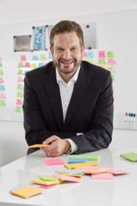 Dr. Christian Ellrich steht an einem Tisch mit bunten Haftnotizen und lächelt in die Kamera.