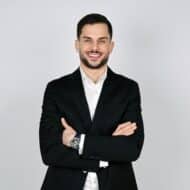 Mann in schwarzem Anzug mit verschränkten Armen und lächelndem Gesicht vor neutralem Hintergrund.