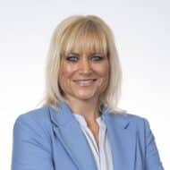 Porträt einer lächelnden Frau mit blonden Haaren, die einen blauen Blazer trägt, vor einem hellen Hintergrund.