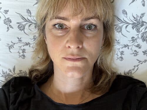 Frau mit blonden Haaren liegt im Bett, schaut direkt in die Kamera, umgeben von einem floral gemusterten Kissen.