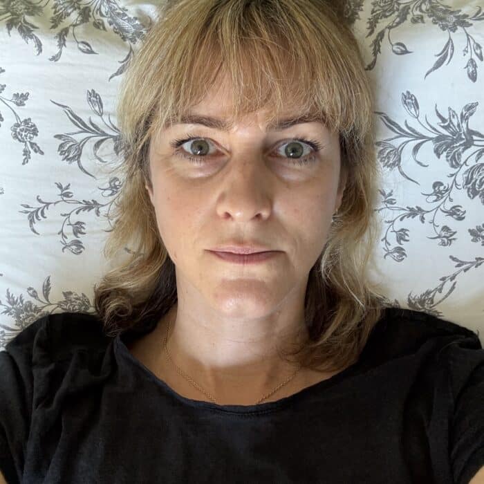 Frau mit blonden Haaren liegt im Bett, schaut direkt in die Kamera, umgeben von einem floral gemusterten Kissen.