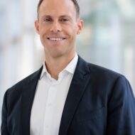 Porträt eines lächelnden Mannes in einem dunklen Blazer und weißem Hemd, vor unscharfem, modernem Hintergrund.