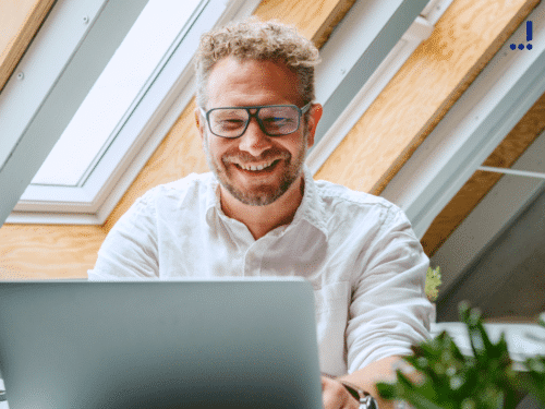 Lächelnder Mann mit Brille sitzt an einem Laptop unter einem Dachfenster, umgeben von Pflanzen.