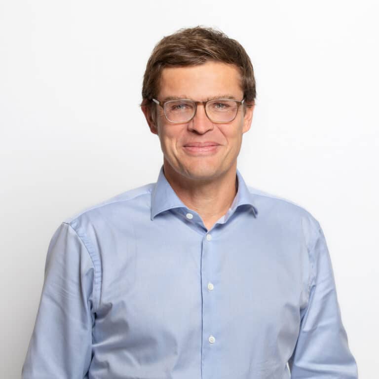 Porträt eines Mannes mit Brille, braunen Haaren, in einem hellblauen Hemd, vor einem weißen Hintergrund.