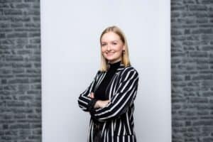 Junge Frau mit blonden Haaren in gestreiftem Blazer, lächelt und steht vor einer hellen Wand mit Steinmuster.