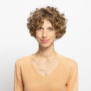 undconsorten Porträt einer lächelnden Frau mit lockigem, blondem Haar, die ein orangefarbenes Oberteil trägt. Hintergrund ist weiß.
