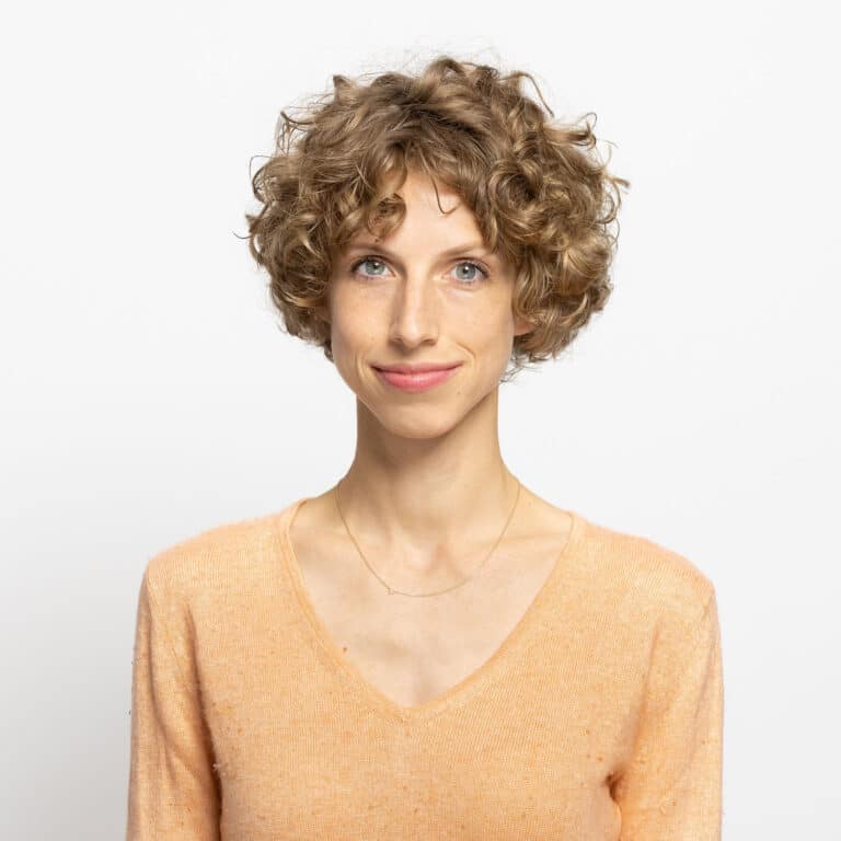 Porträt einer lächelnden Frau mit lockigem, blondem Haar, die ein orangefarbenes Oberteil trägt. Hintergrund ist weiß.