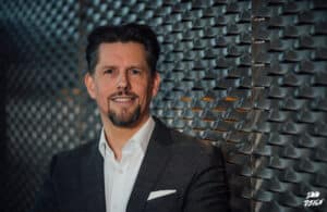 Markus Kamps-1 c Martin Chabinski Mann in Anzug lächelt vor einer strukturierten Wand aus Metallplatten.