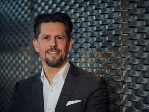 Mann in Anzug lächelt vor einer strukturierten Wand aus Metallplatten.