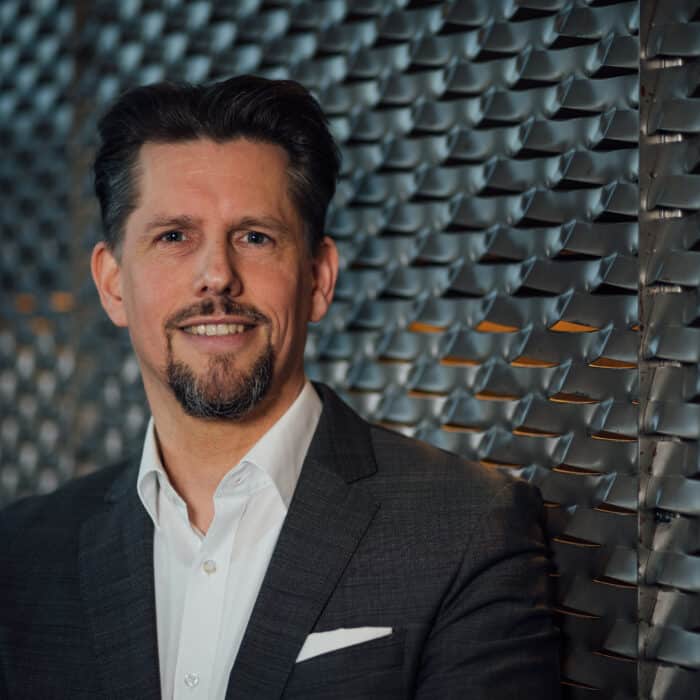 Mann in Anzug lächelt vor einer strukturierten Wand aus Metallplatten.