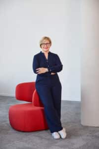 Frau mit blonden Haaren und Brille steht lächelnd vor einer roten Sitzgelegenheit in einem modernen Raum.