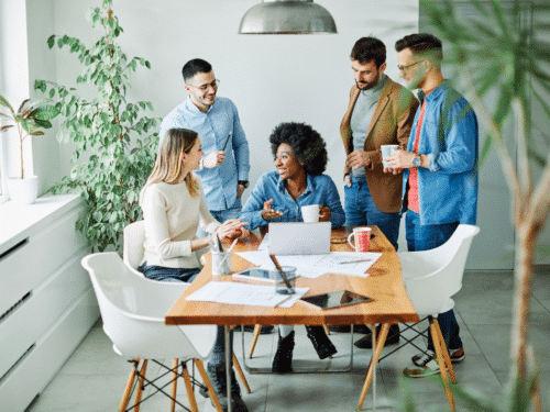 Sechs Personen in einem modernen Raum sitzen um einen Tisch und diskutieren, während Pflanzen im Hintergrund stehen.