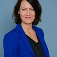 Porträt einer Frau mit schulterlangen, braunen Haaren, die einen blauen Blazer und ein schwarzes Oberteil trägt, vor ein...