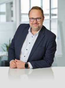 SchmidtJanMartin Mann mit Brille in Anzug steht an einem Tisch, lächelt und posiert vor einem modernen Bürohintergrund.