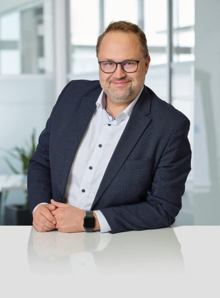Mann mit Brille in Anzug steht an einem Tisch, lächelt und posiert vor einem modernen Bürohintergrund.