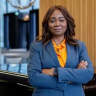 Yvonne Bettkober in einem blauen Anzug mit verschränkten Armen, lächelt vor modernem Hintergrund.