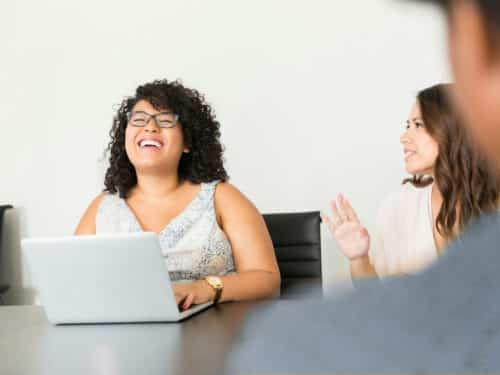 Lächelnde Frau mit Locken sitzt am Tisch mit Laptop, während eine andere Frau im Hintergrund spricht.