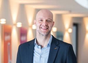 Mann mit Glatze und Bart, trägt ein gestreiftes Hemd und einen Blazer, lächelt vor einem hellen, modernen Hintergrund.