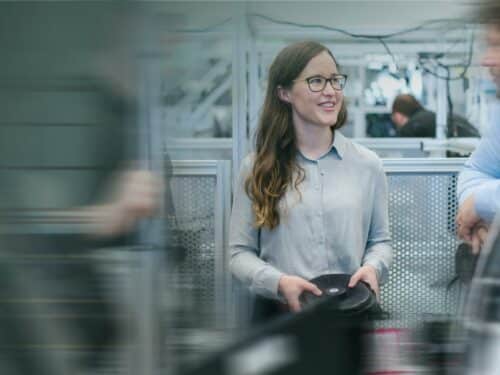 Frau mit Brille hält eine schwarze Platte, lächelt und steht in einem modernen Arbeitsumfeld mit technischen Geräten.
