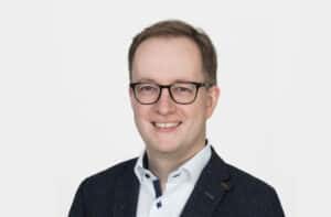 Porträt eines lächelnden Mannes mit Brille, in einem dunklen Blazer und hellblauem Hemd, vor hellem Hintergrund.