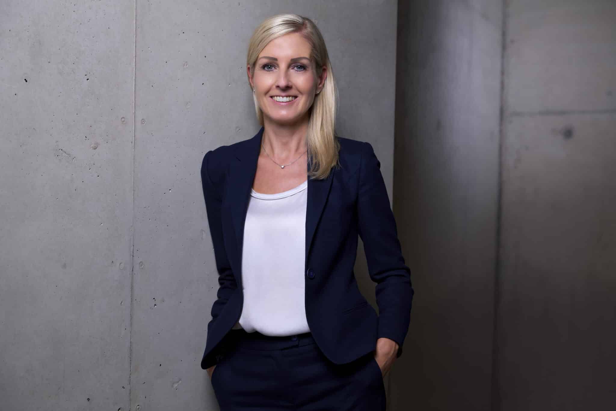 Porträt einer lächelnden Frau in formeller Kleidung, die an einer Betonwand steht.