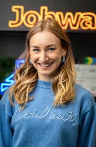 Lächelnde Frau mit blonden Haaren in blauer Sweatshirt, vor einem leuchtenden Schriftzug im Hintergrund.