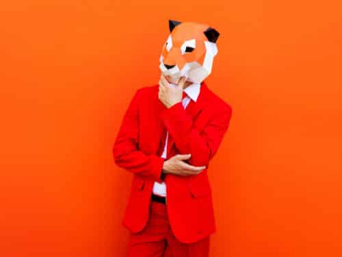 Mann in rotem Anzug mit 3D-Oriami-Tiermaske vor orangefarbenem Hintergrund, nachdenkliche Pose.