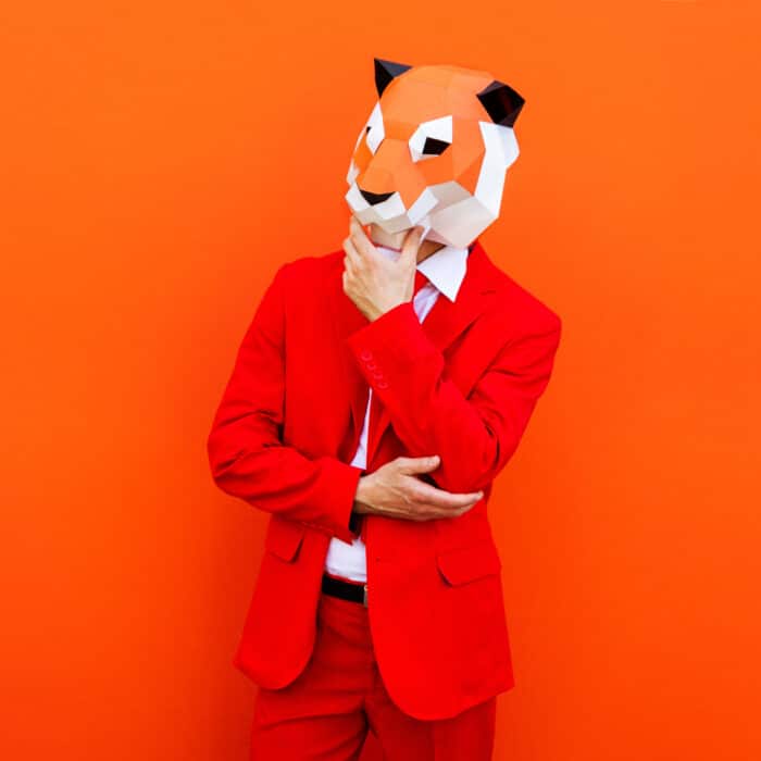 Mann in rotem Anzug mit 3D-Oriami-Tiermaske vor orangefarbenem Hintergrund, nachdenkliche Pose.
