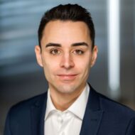 Porträt eines Mannes mit dunklen Haaren, in Anzug und Hemd, vor unscharfem Hintergrund.