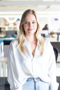 Junge Frau mit blonden Haaren in weißem Hemd steht in einem modernen Büro.