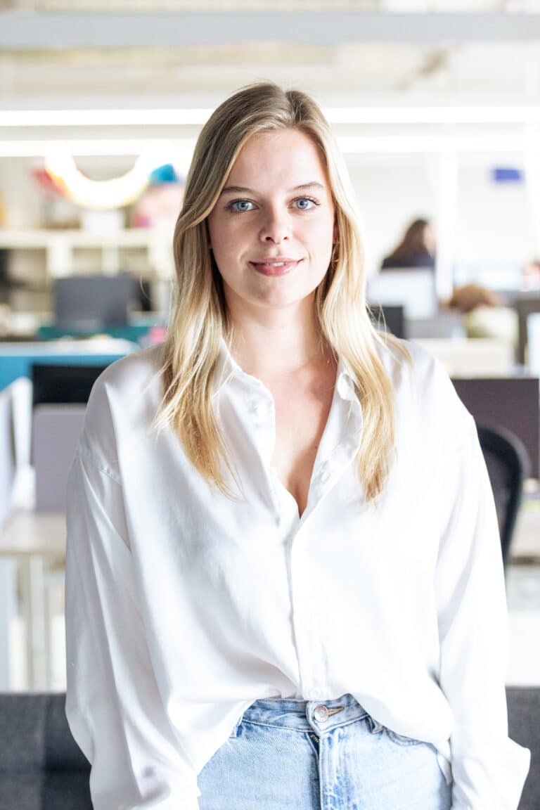 Junge Frau mit blonden Haaren in weißem Hemd steht in einem modernen Büro.