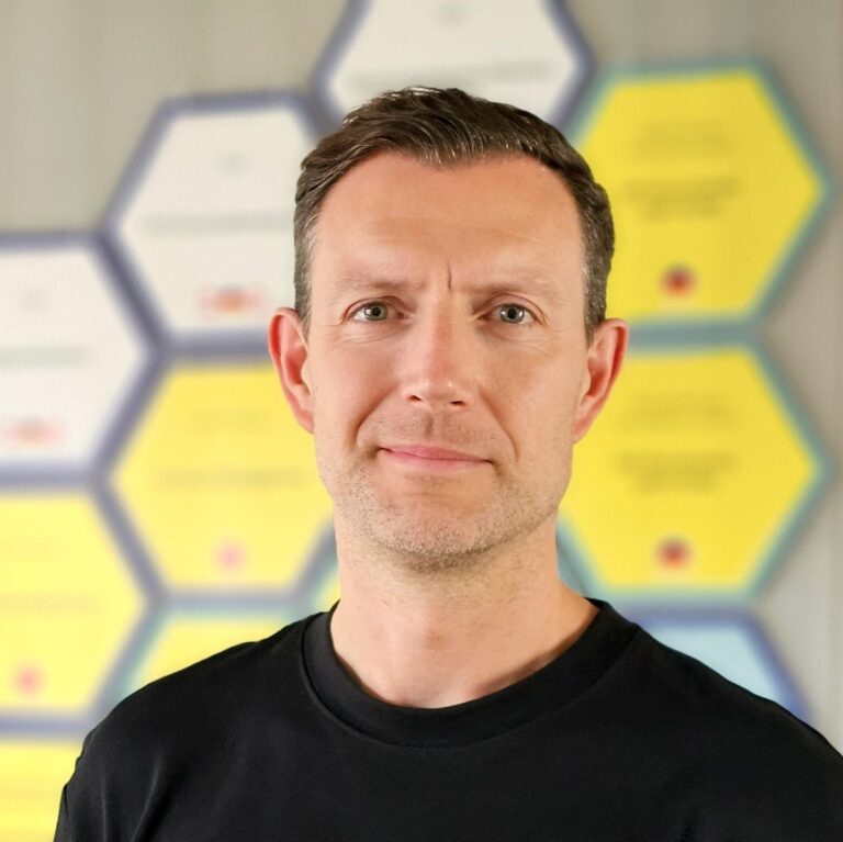 Mann mit kurzem Haar in schwarzem T-Shirt vor einem Hintergrund aus bunten, sechseckigen Plakaten.