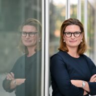 Dr. Charlotte Beissel steht mit verschränkten Armen vor einer Glaswand und lächelt, ihre Reflexion ist sichtbar.