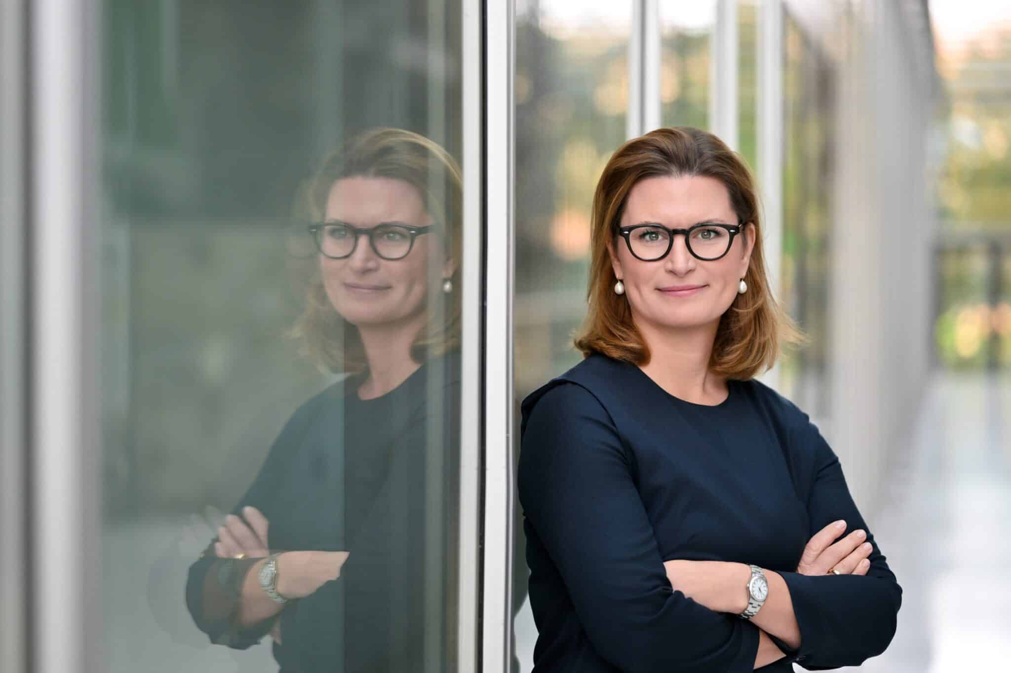 Dr. Charlotte Beissel steht mit verschränkten Armen vor einer Glaswand und lächelt, ihre Reflexion ist sichtbar.