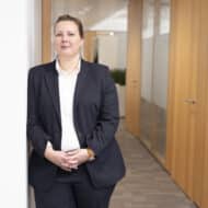 Frau in Anzug steht an einer Wand in einem modernen Büro mit Holzverkleidung und neutralen Farben.