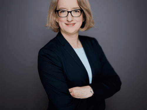 Professionelles Porträt einer Frau mit blonden Haaren, Brille und einem dunklen Blazer, die lächelnd mit verschränkten ...