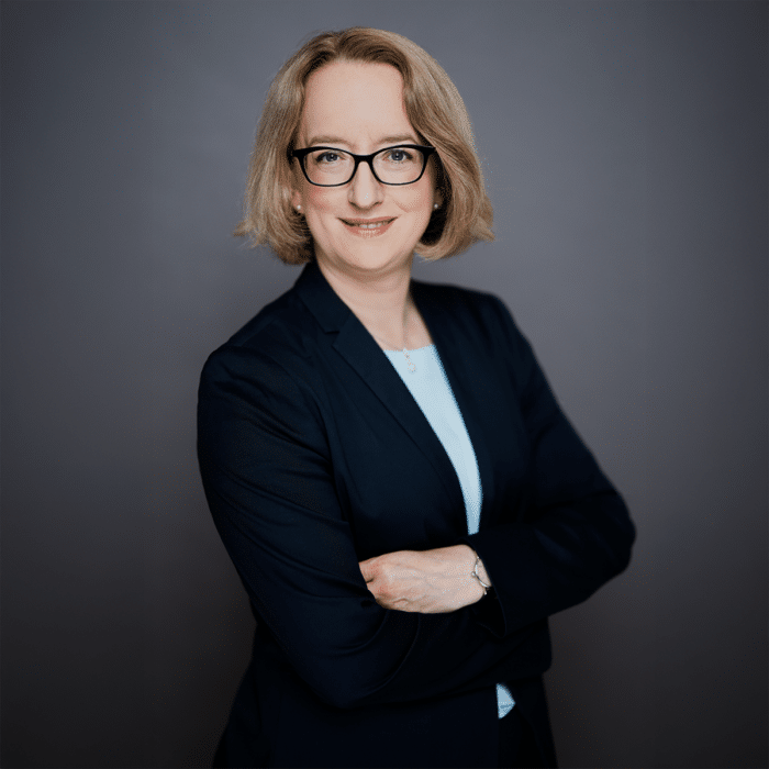 Professionelles Porträt einer Frau mit blonden Haaren, Brille und einem dunklen Blazer, die lächelnd mit verschränkten ...