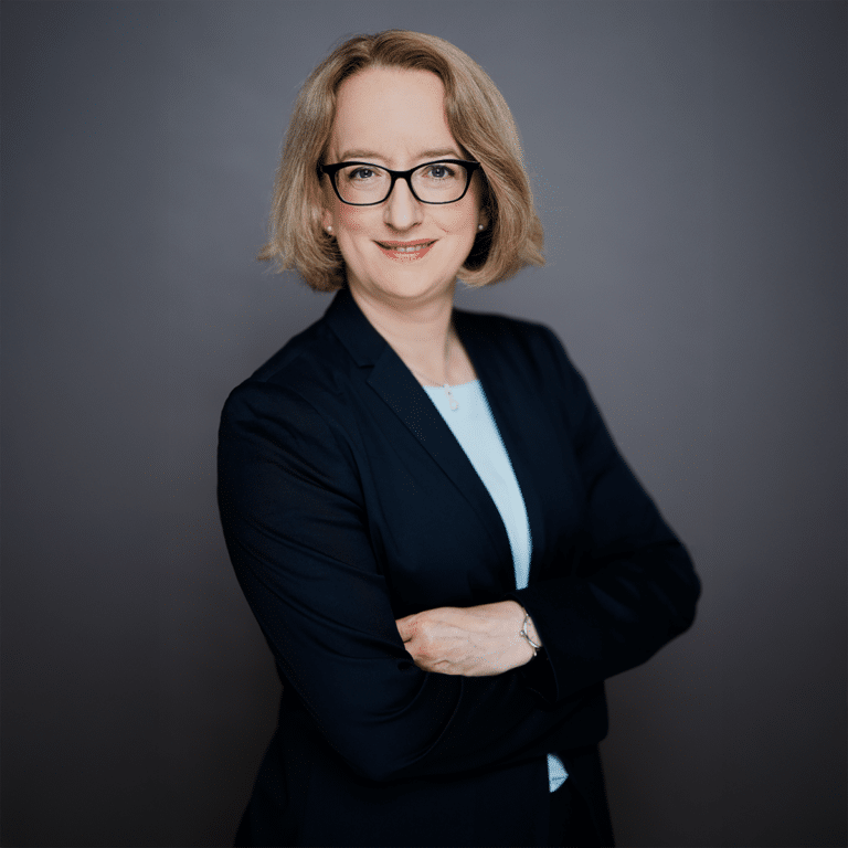 Professionelles Porträt einer Frau mit blonden Haaren, Brille und einem dunklen Blazer, die lächelnd mit verschränkten ...